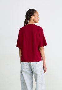 Tricou scurt cu mânecă, de culoare burgundă, cu croială lejeră, din material din bumbac și guler rotund. Este purtat cu pantaloni cargo gri deschis, care au buzunare laterale.