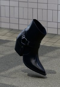 Svart läderankelstövel med spetsig tå, ankelrem, silverdetaljer och klack med flera lager, vilande på en texturerad yta.