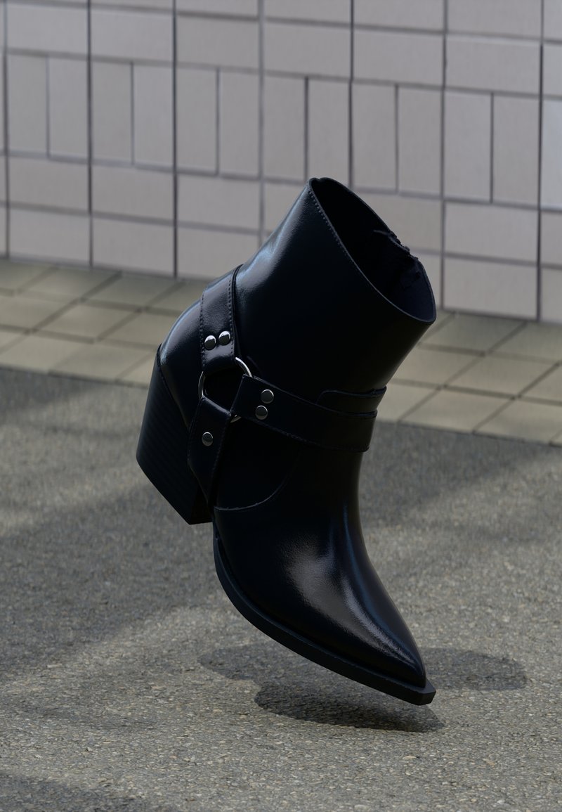 Svart läderankelstövel med spetsig tå, ankelrem, silverdetaljer och klack med flera lager, vilande på en texturerad yta.
