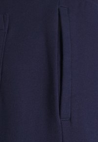 Marinblå joggers i tyg med en mjuk textur, utrustade med sidofickor och söm-detaljer för extra struktur.