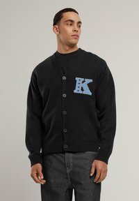 Svart cardigan med V-ringning, fem knappar och en blå "K"-logotyp på vänster bröst. Ribbstickade ärmslut och fåll. Mjukt stickat material.