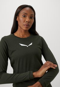 Long-sleeve shirt in dark green fabric with a smooth texture. Features a white angular logo on the chest and a classic round neckline.
