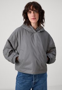 Grå hettejakke laget av lett, glatt materiale med elastiske mansjetter og en frontlomme. Brukt sammen med blå jeans.
