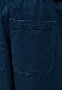 Κοντινό πλάνο σκούρου μπλε παντελονιού από ύφασμα, με τετράγωνη τσέπη με εμφανή ραφή και ελαστική μέση πάνω από αυτήν.