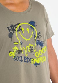 Olive zelené bavlněné tričko s grafikou žlutého usměvavého obličeje, akcenty v námořnické barvě a tučným textem "DOBRÁ ENERGIE" a "KOOL EDICE".