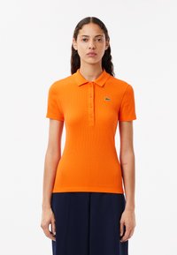 Kortärmad, figurnära polo shirt i klart orange ribbat tyg med krage och treknappars placket; har en grön logotyp som accent.