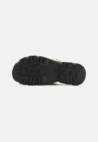 Suela negra de un zapato con un patrón de agarre texturizado y diseño de banda de rodadura, con la parte superior sintética verde parcialmente visible.