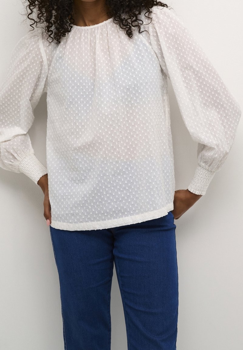 Femme portant un chemisier blanc texturé à manches longues avec des poignets smockés et un jean bleu foncé ajusté, debout les mains sur les hanches.