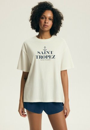 Žena s kučeravými vlasmi, ktorá má na sebe veľké biele tričko s nápisom „Saint Tropez“ a námornícky modré kraťasy, stojaca pred jednofarebným pozadím.