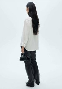 Witte oversized blouse met lange mouwen, gecombineerd met zwarte leren broek. Houdt een kleine zwarte textuurhandtas vast. Minimalistische achtergrond.