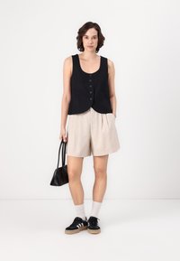 Top sans manches noir avec des boutons, short beige plissé, baskets noires avec des rayures blanches, sac à main noir et chaussettes blanches mi-mollet.