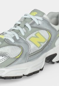 Γκρι και λευκό αθλητικό παπούτσι New Balance με κίτρινο λογότυπο και λευκά κορδόνια, που δείχνει πλευρικές και μπροστινές λεπτομέρειες σε λευκό φόντο.