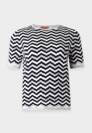 Femme portant un haut à manches courtes noir et blanc à motif ondulé, un pantalon large gris clair et des sandales noires, debout devant un fond uni.