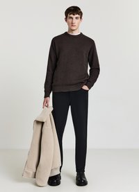 Brauner Strickpullover mit geripptem Saum, schwarze Hose und schwarze Schuhe. Beige Lammfelljacke über den Arm gelegt, glatte Textur, schlichtes Design.