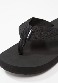 Mustat flip-flopit, joissa on teksturoitu musta nailonihihna ja pehmustettu musta kumipohja. "REEF" -logo näkyy hihnassa.