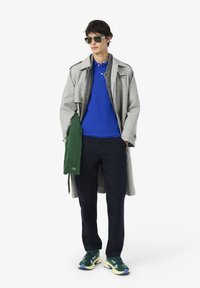 Grå rutig trenchcoat över en blå polo-shirt, grön tröja draperad, marinblå byxor och gröna och blå sneakers med kraftiga sulor.