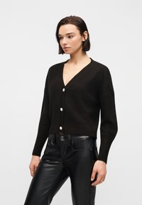 Γυναίκα με κοντά σκούρα μαλλιά που φοράει μαύρη πλεκτή ζακέτα με πέρλες κουμπιά και μαύρο δερμάτινο παντελόνι, όρθια μπροστά σε ανοιχτό φόντο.