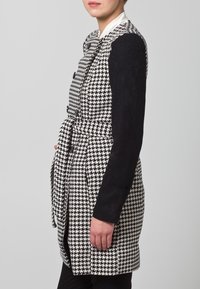 Mustavalkoinen houndstooth-takki, jossa on pitkät mustat hihat, muotoiltu istuvuus ja vyötärönauha. Ominaisuuksiin kuuluvat kaksoispainikkeet ja kaksi taskua.