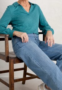 Maglietta a maniche lunghe color turchese con colletto, abbinata a jeans a vita alta blu chiaro, seduta su una sedia di legno. Sfondo neutro.