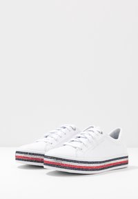 Białe trampki z koronką, z teksturowaną gumową podeszwą, ozdobione paskiem w kolorach czerwonym, granatowym i srebrnym. Górna część wykonana z gładkiej skóry.