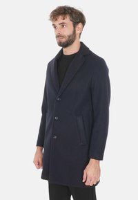 Cappotto in lana navy con rever a taglio, quattro bottoni e tasche laterali. Presenta una vestibilità sartoriale e una texture liscia, ideale per essere indossato a strati.