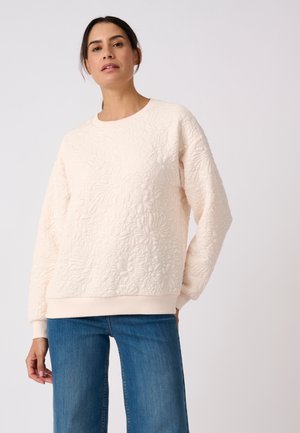 Donna con felpa trapuntata crema a fiori e jeans blu, in piedi con una mano in tasca davanti a uno sfondo chiaro e uniforme.