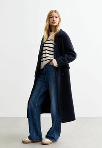Cappotto lungo blu sopra un maglione a righe crema, abbinato a jeans in denim blu a gamba larga e sneakers beige con dettagli bianchi.