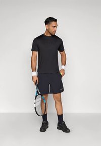 Μαύρο αθλητικό T-shirt με κοντά μανίκια, σε συνδυασμό με ναυτικά σορτς. Κρατά ρακέτα τένις, φορά μαύρα αθλητικά παπούτσια και αντίθετες λευκές περιβραχιόνια.