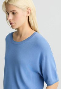 Tricou tricotat de culoare albastru deschis, cu decolteu rotund și mâneci scurte. Țesătura pare moale și netedă, cu o textură subtilă.