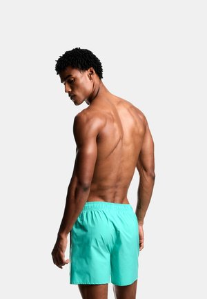 Uomo muscoloso con pelle scura e capelli ricci, visto di spalle, che indossa pantaloncini da bagno turchese brillante su uno sfondo chiaro e uniforme.