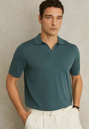 Polo cu mânecă scurtă dintr-un amestec de bumbac de culoare turcoaz, cu guler în V, guler ribbat și textură netedă, asociat cu pantaloni eleganți de culoare deschisă.