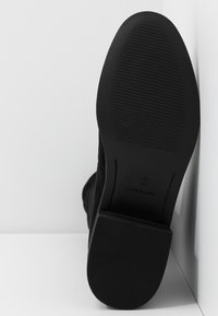 Podeszwa czarnego zamszowego buta z teksturowanym gumowym spodem, rozmiar 5, oparta o białą narożną ścianę i podłogę.