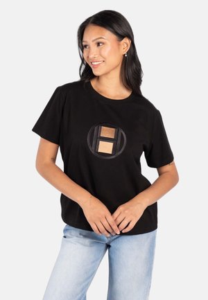 Ung kvinne iført en svart t-skjorte med et rundt merke og lyseblå jeans, smilende og ser til siden.