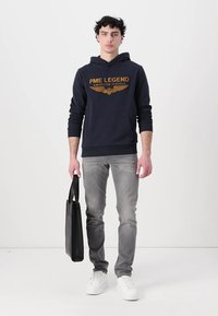 Marineblauwe hoodie met gouden "PME LEGEND AMERICAN CLASSIC" logo, gecombineerd met lichtgrijze jeans en witte sneakers; houdt een zwarte tote bag vast.