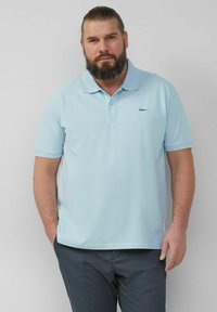 Helles blaues Poloshirt mit weicher Textur, kurzen Ärmeln und Kragen. Mit kleinem Logo auf der linken Brust. Kombiniert mit dunkel gefärbten Hosen.