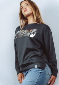 Młoda kobieta z długimi blond włosami, ubraną w ciemną bluzę z odblaskowym napisem "mapalt" oraz jasnoniebieskie dżinsy, na jednolitym tle.