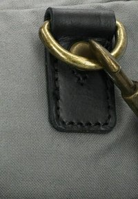 Primer plano de un broche de latón sujeto a un parche de cuero negro cosido sobre un fondo de tejido gris.