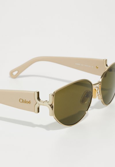 Gafas de sol beige con acentos dorados, lentes ovalados verdes y una bisagra de diseño cruzado distintivo. El material parece suave con un acabado mate.