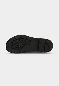 Semelle de chaussure en caoutchouc noir avec des rainures profondes et une adhérence à motifs sur un fond gris clair uni.