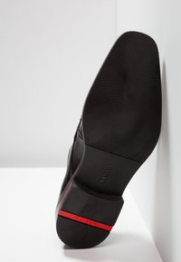 Semelle de chaussure en cuir noir avec caoutchouc texturé, présentant une bande accent rouge et un logo gravé. Le talon est légèrement surélevé.