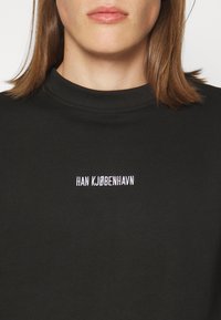Personne aux cheveux bruns mi-longs porte un haut noir à col montant avec un texte blanc "HAN KJØBENHAVN" sur la poitrine.