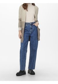 Beige und grauer langer cardigan über einem beigen gerippten Rollkragenpullover, kombiniert mit hochgeschnittenen blauen Jeans und schwarzen klobigen Schuhen.