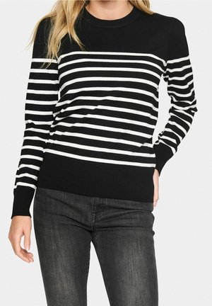 Žena v černobílém horizontálně pruhovaném svetru s dlouhým rukávem a tmavě šedých džínách stojí s jednou rukou v kapse.