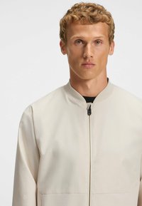 Hellbeige Bomberjacke mit hexagonalem, geprägtem Muster, Reißverschluss vorne, Stehkragen und kontrastierender schwarzer Innenauskleidung. Glatte Stofftextur.