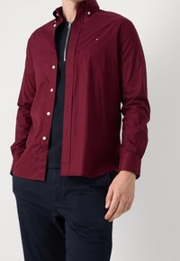 Bordowa koszula z długim rękawem, zapinana na guziki, z kołnierzem typu spread, białymi guzikami i logo na piersi, noszona na granatowej bluzce i czarnych spodniach.