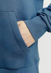 Felpa blu con tessuto strutturato, dotata di tasca frontale e polsini aderenti, che mette in risalto la morbidezza del materiale e i dettagli del design.