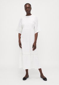 Rochie albă, până la genunchi, cu un model texturat în carouri, margine din dantelă și mâneci largi, completată de pantofi negri fără toc.