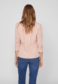 Blazer ajusté rose blush à manches froncées, design structuré et texture lisse. Associé à un jean en denim bleu.