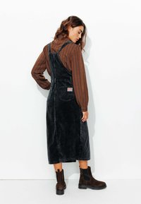 Robe salopette en velours côtelé noir avec une silhouette droite, bretelles ajustables, poche avant et étiquette. Associée à des bottines beige.
