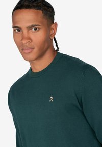 Grüner Strickpullover mit Rundhalsausschnitt, versehen mit einem kleinen goldenen Akzent-Logo auf der linken Brust. Glatte Textur, taillierte Passform.